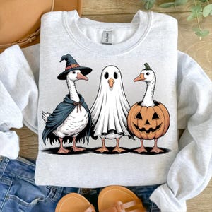 Op de afbeelding: Een lichtgrijs sweatshirt met een Halloween-thema. Het toont drie ganzen verkleed als heks, spook en pompoen. De ganzen zijn afgebeeld met zwarte contouren, wat een cartoonachtig effect cre&euml;ert.
