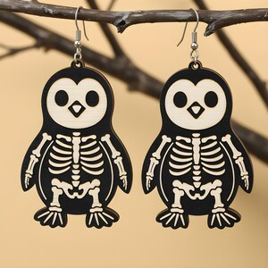 May include: A pair of wooden earrings shaped like penguins with skeleton designs. The earrings are black with natural wood accents and silver-colored ear wires. The penguin's face and skeleton are light-colored, creating a contrast against the black body. The earrings are hanging from a brown branch.