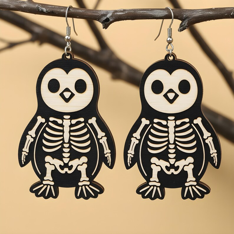 May include: A pair of wooden earrings shaped like penguins with skeleton designs. The earrings are black with natural wood accents and silver-colored ear wires. The penguin's face and skeleton are light-colored, creating a contrast against the black body. The earrings are hanging from a brown branch.