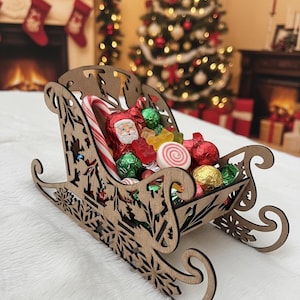 May include: A wooden sleigh filled with Christmas candies, including a candy cane, wrapped chocolates, and gummy candies. The sleigh has decorative cutouts and sits on a white surface. A Christmas tree and fireplace are in the background.