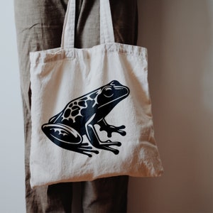 May include: A cream-colored tote bag featuring a large black and white graphic of a frog. The frog design is detailed with spots and outlines. The bag has two handles and is made of a sturdy fabric.