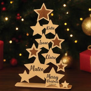 Puede incluir: Adorno de árbol de Navidad de madera en forma de estrellas. El árbol tiene nombres grabados en las estrellas, incluyendo "Katie", "Isaac", "Claire" y "Mateo". La base del árbol dice "Merry Christmas Forever".