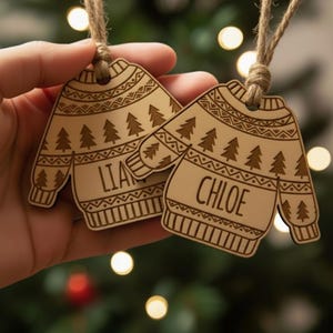 May include: Two wooden Christmas sweater ornaments with the names "LIA" and "CHLOE" engraved on them. The ornaments are light brown with a sweater design, including trees and decorative patterns. Each ornament has a jute string for hanging.