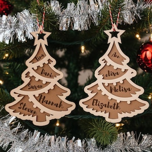 May include: Two wooden Christmas tree ornaments with names engraved on them. The ornaments are light brown with a darker brown interior. Each tree has a star at the top and is hanging from a red and white string. The names on the ornaments are Lira, Dari, Susan, Hannah, Steven, Ann, elisa, Melisa, and Elizabeth.