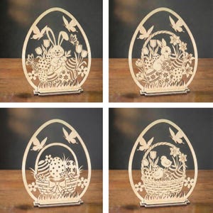 Könnte beinhalten: Vier hölzerne Ostereier-Dekorationen. Jeder eiförmige Ausschnitt zeigt ein anderes Design: ein Hase mit Eiern, ein Hase in einem Korb, ein Korb mit Eiern und ein Korb mit Küken. Die Ausschnitte umfassen Blumen, Schmetterlinge und Vögel.
