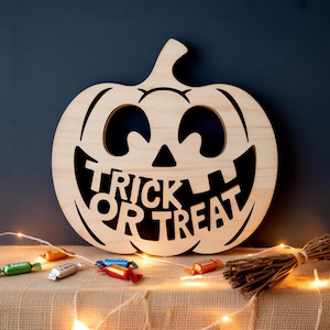 Puede incluir: Decoración de Halloween con forma de calabaza de madera con las palabras "Trick or Treat" recortadas. La calabaza tiene una cara sonriente con ojos y boca recortados. Caramelos y una escoba en primer plano.