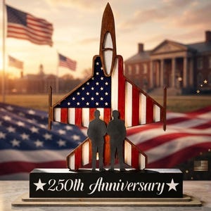 May include: A wooden jet-shaped sculpture with the American flag design on the wings, and two silhouetted figures standing in front. The base is black with the text "250th Anniversary" in white. American flags wave in the background.