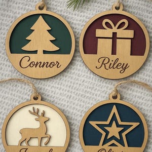 May include: Four wooden Christmas ornaments with different designs and names: a tree with "Connor", a gift with "Riley", a deer with "Joseph", and a star with "Olivia". Each ornament has a natural wood finish, a colored background, and a jute string for hanging.