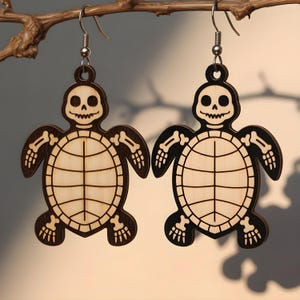 May include: A pair of wooden earrings featuring a skeleton turtle design. The earrings are laser-cut with a light wood center and dark brown outline, including the turtle's shell, head, and limbs. The earrings hang from silver-colored fish hook ear wires.