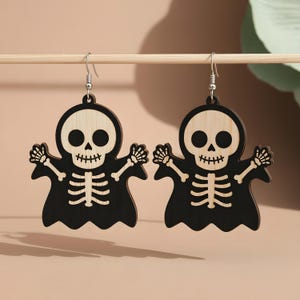 May include: A pair of Halloween-themed earrings. Each earring features a black skeleton design with a light wood-colored skull and ribcage. The earrings are hanging from silver-colored hooks. The skeleton is wearing a black cloak.