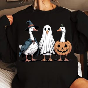 Op de afbeelding: Zwart sweatshirt met een Halloween-thema, met drie ganzen in kostuums. E&eacute;n is een heks, een andere een spook en de derde zit in een pompoen. De ganzen zijn wit met oranje snavels en poten.