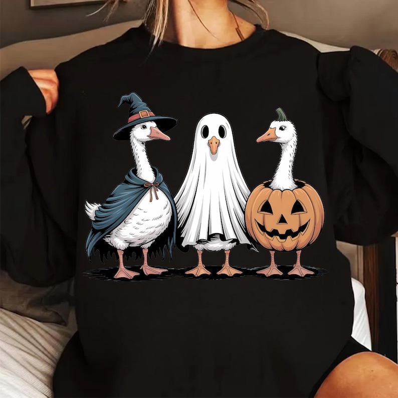 Op de afbeelding: Zwart sweatshirt met een Halloween-thema, met drie ganzen in kostuums. E&eacute;n is een heks, een andere een spook en de derde zit in een pompoen. De ganzen zijn wit met oranje snavels en poten.