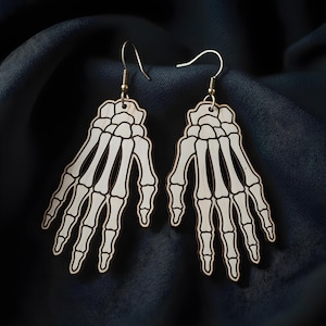 Puede incluir: Un par de pendientes de mano de esqueleto. Los pendientes están hechos de madera de color claro con un diseño detallado de hueso blanco y contornos oscuros. Cada pendiente tiene un gancho dorado para orejas perforadas. El fondo es una tela oscura y texturizada.