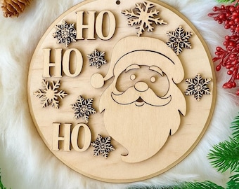 May include: A wooden Christmas ornament featuring a Santa Claus face, the words "HO HO HO", and snowflake accents. The ornament is round with a hole at the top for hanging. The design is laser cut from light-colored wood.