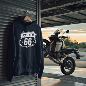May include: A black hoodie with a white Route 66 sign graphic on the front. The hoodie is hanging on a hanger in front of a motorcycle.
