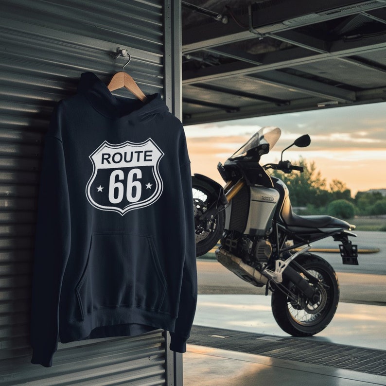 May include: A black hoodie with a white Route 66 sign graphic on the front. The hoodie is hanging on a hanger in front of a motorcycle.