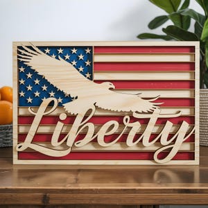 May include: Wooden American flag with a laser-cut bald eagle and the word "Liberty". The flag features red and white stripes, a blue field with white stars, and a natural wood eagle and lettering. The piece is framed.