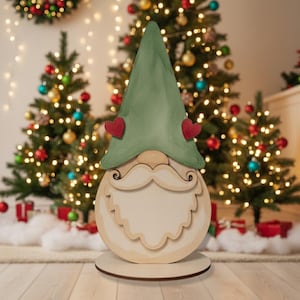 Könnte beinhalten: Weihnachtliche Wichtel-Dekoration aus Holz mit grünem Hut, roten Herzakzenten und weißem Bart. Der Wichtel steht auf einem runden Holzsockel. Weihnachtsbaumschmuck im Hintergrund.