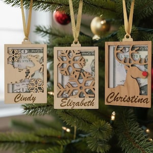 May include: Three wooden Christmas ornaments hanging on a tree. Each ornament has a rectangular frame with a decorative cutout design and a name. The ornaments are filled with money and have gold ribbon hangers.