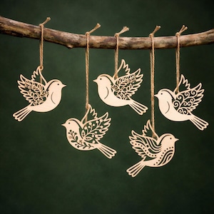 Może przedstawiać: Pięć drewnianych ozdób w kształcie ptaków zawieszonych na gałęzi za pomocą sznurka. Każdy ptak ma unikalny ażurowy wzór na skrzydłach i ogonie. Ptaki są jasne na ciemnozielonym tle, tworząc naturalną, rustykalną estetykę.