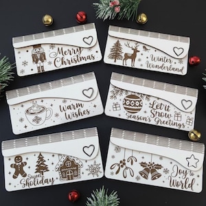 May include: A collection of six wooden Christmas-themed gift card holders. Each holder features a unique design, including a nutcracker, deer, gingerbread house, and festive text like "Merry Christmas" and "Winter Wonderland". The holders have a heart-shaped clasp.