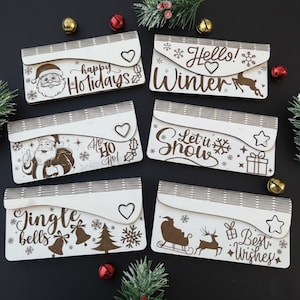 Op de afbeelding: Zes houten cadeaubonnenhouders met kerstthema. Elke houder heeft een uniek ontwerp met feestelijke teksten zoals "Vrolijk Kerstfeest" en illustraties van de kerstman, sneeuwvlokken en rendieren.