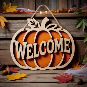 以下が含まれることがあります： 「WELCOME」の文字がくり抜かれた、木製のカボチャ型のサイン。オレンジ色の背景と天然木のフレームがあります。ロープで吊り下げられており、秋の葉やドングリに囲まれています。