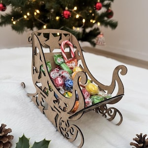 May include: A wooden sleigh filled with colorful wrapped candies and a candy cane. The sleigh has a decorative star and swirl design. The background includes a Christmas tree with red ornaments and lights.