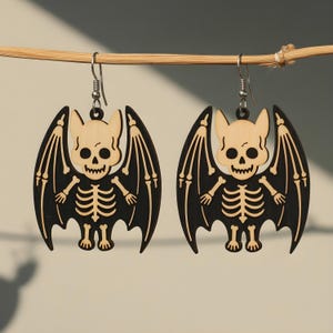 May include: A pair of Halloween-themed earrings. Each earring features a bat design with a skeleton body and wings. The bat's head and skeleton are light-colored, while the wings are black. The earrings are suspended from silver-colored hooks.
