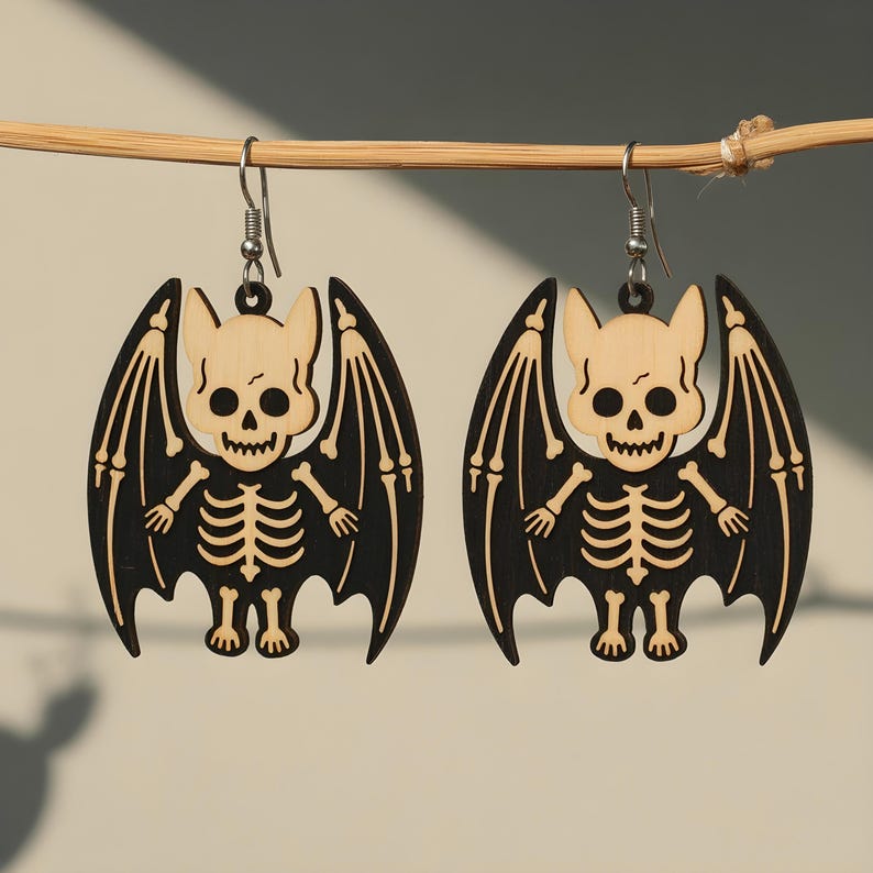 May include: A pair of Halloween-themed earrings. Each earring features a bat design with a skeleton body and wings. The bat's head and skeleton are light-colored, while the wings are black. The earrings are suspended from silver-colored hooks.