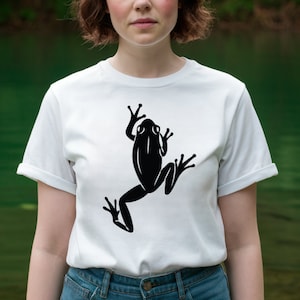May include: White t-shirt featuring a large black frog graphic. The frog is depicted in a stylized, minimalist design. The shirt has short sleeves and a crew neck. The model is wearing blue jeans.