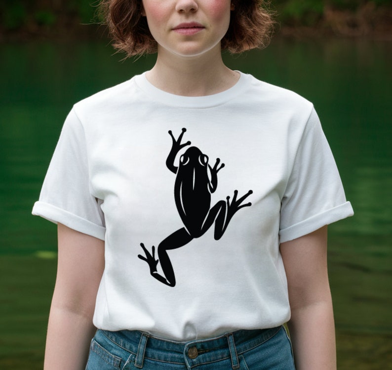 May include: White t-shirt featuring a large black frog graphic. The frog is depicted in a stylized, minimalist design. The shirt has short sleeves and a crew neck. The model is wearing blue jeans.