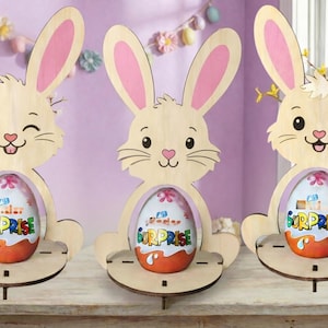 Op de afbeelding: Drie houten paashaasdecoraties met roze binnenoren en een lachend gezicht. Elke haas houdt een Kinder Surprise-ei vast. De achtergrond bevat paasversieringen en een raam met gele bloemen.