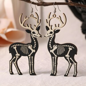 May include: A pair of wooden earrings featuring a deer skeleton design. The earrings are black with white bone details and silver-colored ear wires. The deer have large antlers and are in a standing pose. The earrings are a unique accessory.
