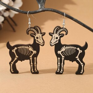 May include: A pair of black goat earrings with a skeleton design. The earrings are made of wood and feature a detailed depiction of a goat's skeleton. The earrings have silver-colored hooks.