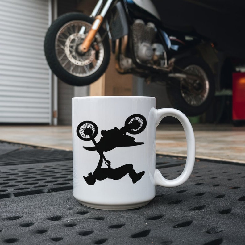 May include: White ceramic mug with a black silhouette of a person doing a backflip on a dirt bike. The mug has a handle on the right side.