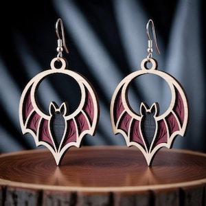 May include: Wooden bat earrings with a layered design. The earrings feature a bat silhouette within a circular frame, with burgundy and black accents. The earrings hang from silver-colored ear wires.