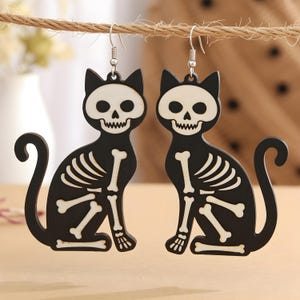 May include: A pair of black cat-shaped earrings with white skeleton designs. The earrings are hanging from silver hooks and feature a sitting cat silhouette with bone details, perfect for Halloween.