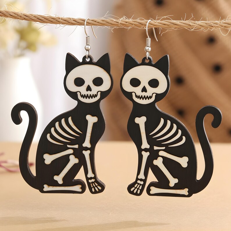May include: A pair of black cat-shaped earrings with white skeleton designs. The earrings are hanging from silver hooks and feature a sitting cat silhouette with bone details, perfect for Halloween.