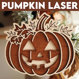 Puede incluir: Un adorno de calabaza de madera con una cara de calabaza tallada y hojas en la parte superior. La calabaza es marrón y la cara está recortada para revelar la madera clara de abajo.