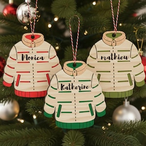 Peut inclure: Trois décorations de Noël en bois en forme de manteaux d'hiver, chacune avec un nom : "Monica", "Katherine" et "Mathieu". Les manteaux sont de couleur crème avec des accents rouges et verts, suspendus à un sapin de Noël.