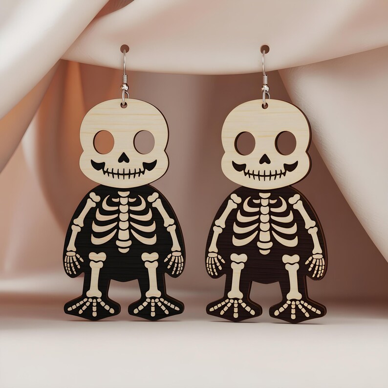 May include: A pair of Halloween-themed earrings featuring a skeleton design. The earrings are made of wood, with a light-colored head and a black body. The skeleton's details are etched into the wood, and the earrings hang from silver-colored hooks.