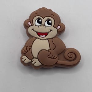 May include: A brown cartoon monkey charm with big eyes and a smiling mouth. The monkey is sitting with its arms crossed and has a curled tail.