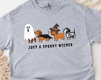 Puede incluir: Camiseta gris claro con un diseño temático de Halloween. El diseño incluye un fantasma, tres perros salchicha disfrazados y el texto "JUST A SPOOKY WIENER". Los perros salchicha están disfrazados de bruja, esqueleto y fantasma.