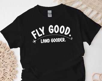 May include: Black t-shirt with white text that reads "FLY GOOD. LAND GOODER." with a small airplane icon.