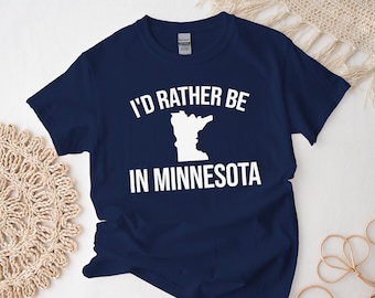 May include: A navy blue t-shirt with white text that reads "I'd rather be in Minnesota" and a white outline of the state of Minnesota.