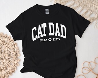 May include: Black t-shirt with white text that reads "Cat Dad" with the names "Bella" and "Kitty" underneath. A paw print is between the names.