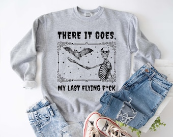 May include: A light gray sweatshirt with a black graphic of a skeleton holding a bat, with the text "THERE IT GOES, MY LAST FLYING F*CK". The sweatshirt is paired with blue jeans and white sneakers.