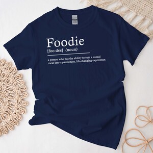 Può includere: Una maglietta blu navy con testo bianco che recita "Foodie [foo-dee] (sostantivo) una persona che ha la capacità di trasformare un pasto informale in un'esperienza appassionante e che cambia la vita."