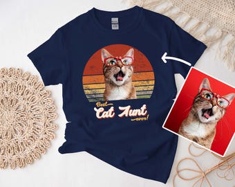 Pode incluir: Camiseta azul marinho com um gato usando óculos vermelhos, em um design retrô de pôr do sol. O texto "Best Cat Aunt ever!" está abaixo da imagem do gato. Uma imagem menor do gato está à direita.